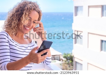 Lovery mature blonde lady having fun conversation with friends standing outdoor with blue sea on background.  Beautiful caucasian curly woman using mobile phone standing on balcony.