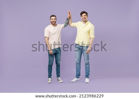Full size length two young smiling happy men friends together in casual t-shirt meeting together greeting give high five clapping hands folded isolated on purple background studio Tattoo translate fun Royalty-Free Stock Photo #2123986229