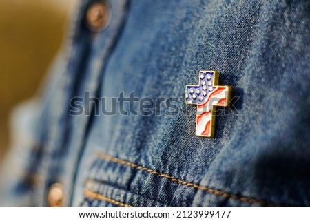 Close up of Christian cross pin with American flag colors is pinned on blue jeans jacket. Patriotism and religious rights concept