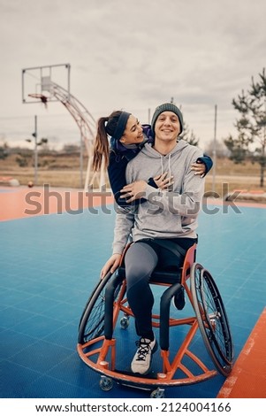 Happy athletic woman embracing her male friend with disability who is using wheelchair on basketball court. 