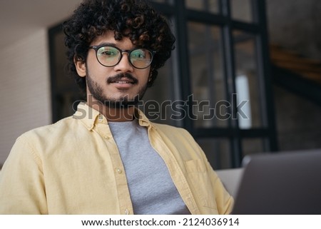 Close up portrait of confident Indian  IT specialist using laptop computer working from home. Young stylish asian businessman wearing eyeglasses sitting in modern office. Successful business concept 