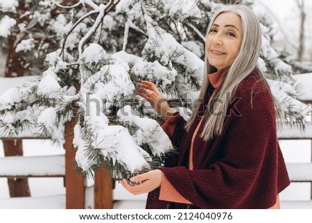 Senior woman with gray hair in a knitted sweater and scarf, sincerely smiling holding a Christmas tree branch on a winter background.