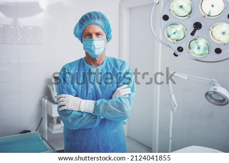Handsome surgeon in surgical wear is looking at camera, standing with crossed arms in a modern operating room