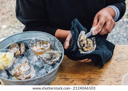 woman shucking oyster at table outdoors with ice-filled tub of oysters and condiments Royalty-Free Stock Photo #2124066503