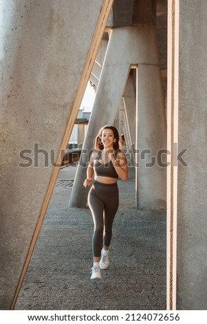 Young multi cultural woman smiling to camera while jogging on urban city to lose weight before summer to get a defined body. Running and workout outdoors. Sunset scene with young athletes