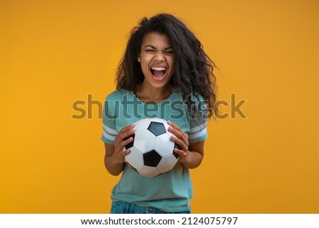 Studio image of excited soccer fan girl holding ball in hands looking at the camera with amazed face expression, celebrating her favorite team victory. Isolated over yellow background.