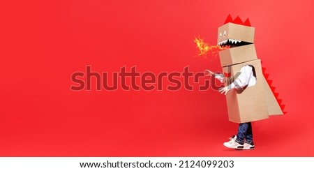 Childhood dreams. Funny little boy plays a cardboard dragon breathing fire and walking on a red background. Fantasy, imagination. Full-length studio portrait. Copy space.