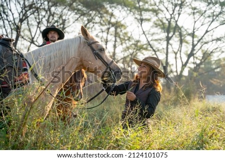 Pretty young Asian woman with cowboy costume smile and stand in front of white horse and stay near other cowboy man in field near village.