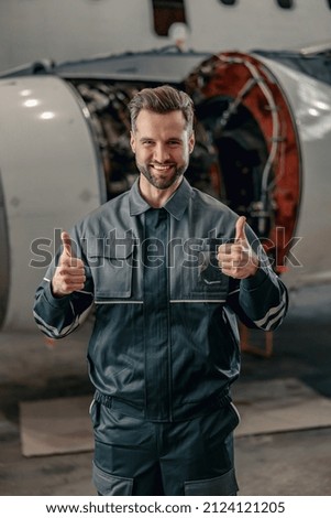 Joyful man aircraft maintenance engineer giving thumbs up and smiling while standing near airplane Royalty-Free Stock Photo #2124121205