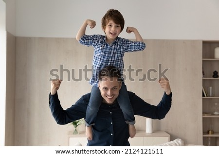 Cheerful little boy riding on happy dads neck and shoulders, making powerful hands. Strong daddy keeping balance with kid on top, flexing arm muscles, smiling at camera. Father and son portrait Royalty-Free Stock Photo #2124121511
