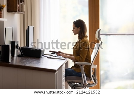 Concentrated successful young Caucasian businesswoman working on online project on computer in modern home workplace, sitting in comfortable office armchair, communicating distantly or web surfing. Royalty-Free Stock Photo #2124121532