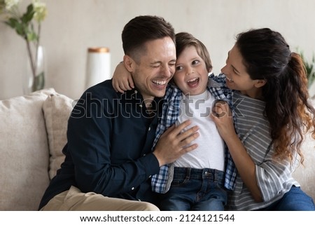 Happy young couple of parents cuddling and tickling adorable little son on couch, laughing, having fun together. Excited mom, dad, kid playing at home, feeling joy, giggling. Family time Royalty-Free Stock Photo #2124121544