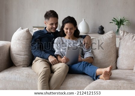 Millennial husband and wife using online application on tablet, watching media content, movie, talking on video call together. Happy couple holding digital gadget, relaxing on cozy couch at home, Royalty-Free Stock Photo #2124121553