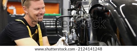 Smiling locksmith with wrench near motorcycle wheel in car workshop. Motorcycle warranty service concept