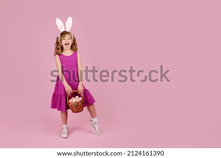 Full body little beautiful smiling girl in Easter bunny ears holds a basket with eggs on pink background. Copy space