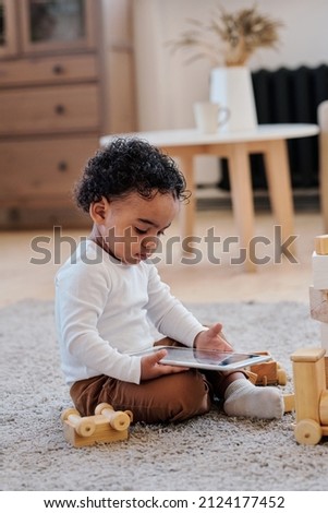 Concentrated curly black boy sitting on carpet in living room and watching cartoons on tablet