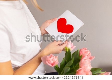 Spring flowers and greeting card. Happy mom holds a card with a red heart in her hands