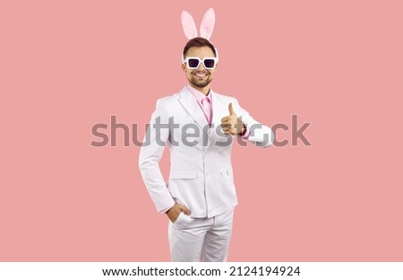 Handsome man with bunny ears wishing you Happy Easter. Happy attractive young guy in white stylish classic suit and funny plush rabbit ears standing isolated on solid pastel pink and showing thumbs up