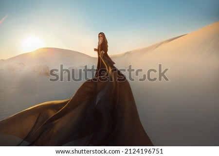 Mystery arabic woman in black long dress stands in desert long train silk fabric fly flytter in wind motion. clothes gold accessories hide face. Oriental fashion model. Sand dunes background sunset Royalty-Free Stock Photo #2124196751