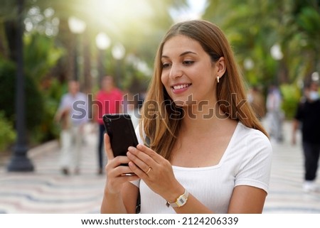 Excited young woman watching her smartphone when walking in the street with blurred people on the background. Beautiful girl using mobile app outdoors. Teenager lifestyle technology concept.