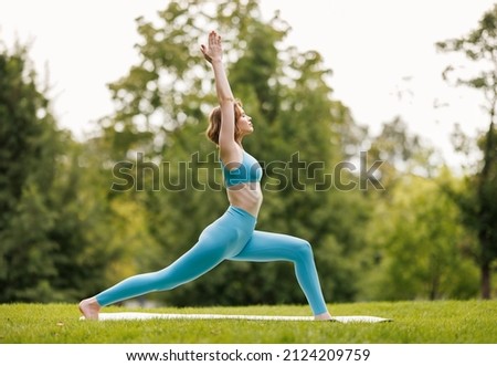 Mental and physical health. Slender focused girl in sportswear performing yoga outside in early morning in nature, female standing in warrior   pose with hands raised on grass in city park Royalty-Free Stock Photo #2124209759