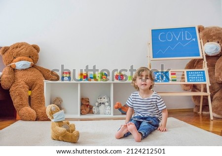Cute little child using a smartphone while sitting on the floor surrounded by toys wearing face masks, during Coronavirus COVID 19 pandemic. Digital device and screen time addiction concept