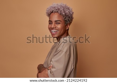 Young overjoyed African American woman laughing with arms folded in front of him rejoicing at news or meeting with friends or boyfriend stands on brown background in studio. communication, people