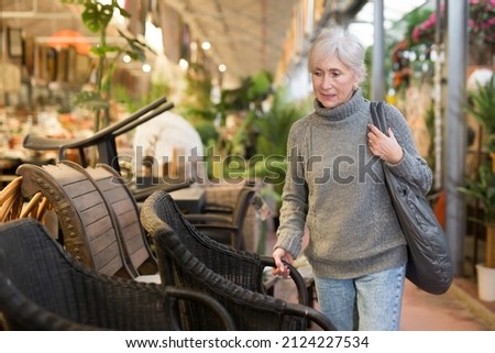 Interested elderly woman visiting furnishing store looking for practical plastic wicker chairs for country house patio ..