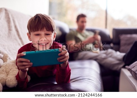Father Uses Mobile Phone As Son Plays Computer Game On Portable Gaming Device At Home In Pyjamas Royalty-Free Stock Photo #2124244166