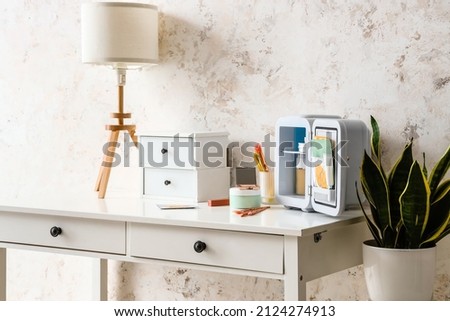 Open cosmetic refrigerator with products, drawers and lamp on table near light wall