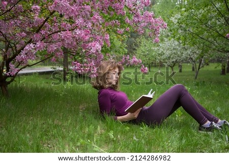 young woman, student, reads book with interest, lying under tree on spring day in park. girls - teenager 17 years old with book on grass. preparation for exams. Spring leisure. Passion for reading Royalty-Free Stock Photo #2124286982