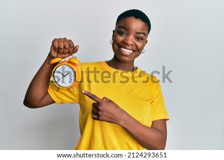 Young african american woman holding alarm clock smiling happy pointing with hand and finger  Royalty-Free Stock Photo #2124293651