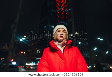 Attractive woman in a red jacket stands at night on the street against the backdrop of modern illuminated architecture and looks away.