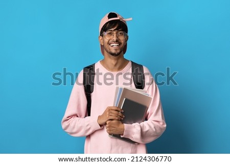 Positive millennial indian guy student in pink baseball cap and sweatshirt posing on blue background, holding books and notepads, carrying backpack, copy space. Education, school, college concept Royalty-Free Stock Photo #2124306770