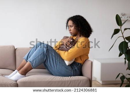Unhappy young black woman suffering from depression, hugging pillow, sitting on couch at home. Upset African American lady feeling hopeless, having mental illness or post traumatic stress disorder Royalty-Free Stock Photo #2124306881