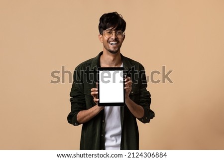 Emotional handsome hindu guy wearing eyeglasses showing brand new digital tablet with white empty screen and smiling, recommending nice online offer, mockup, beige studio background