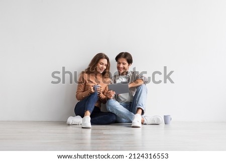 Satisfied millennial caucasian family planning future home interior with tablet, sit on floor with coffee on white wall background in empty room. Moving together, buy and rent apartment with app