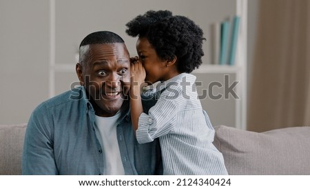 Little african american kid girl shares secret with dad tells amazing fun story in ear funny father smiling listening to cute daughter sitting on sofa trusting relationship closeness parent and child Royalty-Free Stock Photo #2124340424