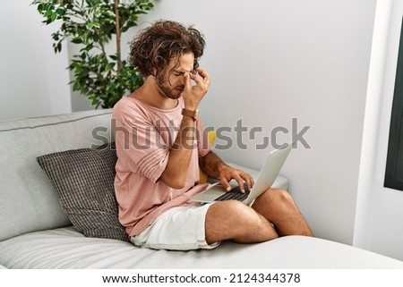 Young hispanic man sitting on the sofa at home using laptop tired rubbing nose and eyes feeling fatigue and headache. stress and frustration concept. 