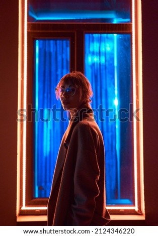 Cyberpunk girl in stylish jacket wears eyeglasses looking to camera, standing against window frame with neon lights at dark street