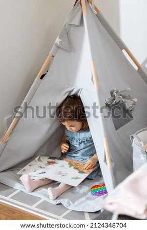 childhood, hygge and people concept - little baby girl reading picture book in kid's tent or teepee at home