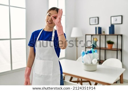 Young chinese girl wearing cleaner uniform standing at home smiling happy doing ok sign with hand on eye looking through fingers 