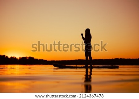Caucasian woman pictured from low angle on paddle board with oar in hands looking at beautiful sunset covering water surface with orange color in evening. Active lifestyle.