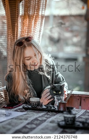 A girl is sitting in a cafe and holding a black cat in her arms, who is trying to drink from a cup