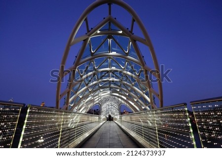 Panorama of the Bridge of Peace, a pedestrian bridge made of steel and glass with a curvy design, over the Kura mtkvari river in the city of Tbilisi, Georgia. Royalty-Free Stock Photo #2124373937