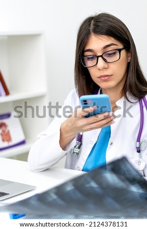 A young dark-haired female doctor with glasses examines an MRI scan in a medical office and takes a picture on her smartphone.Medical concept.