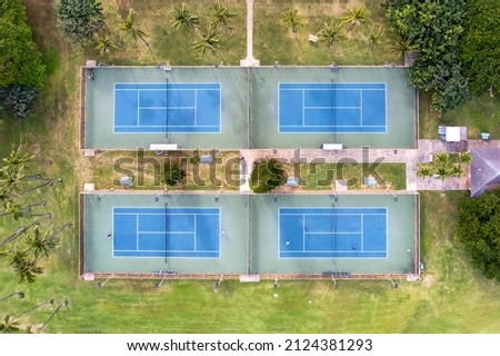 Direct overhead view of four tennis courts in a park surrounded by palm trees and others