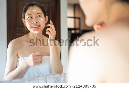 Asian woman smiling while looking her face in the mirror after applying moisturizer cream on her face for solve skin problem.