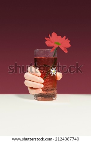 Shot of a female hand holding a glass with a drink with a picture of white flowers and a decorative red flower. The glass is on the light surface. The tableware is on the burgundy background.