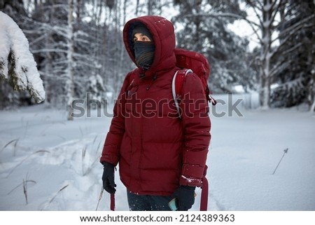 Picture of male hiker in red winter coat with hood wearing black snood scarf up to his nose and gloves, with backpack, standing in the middle of snowy forest, lost, holding smartphone in hands